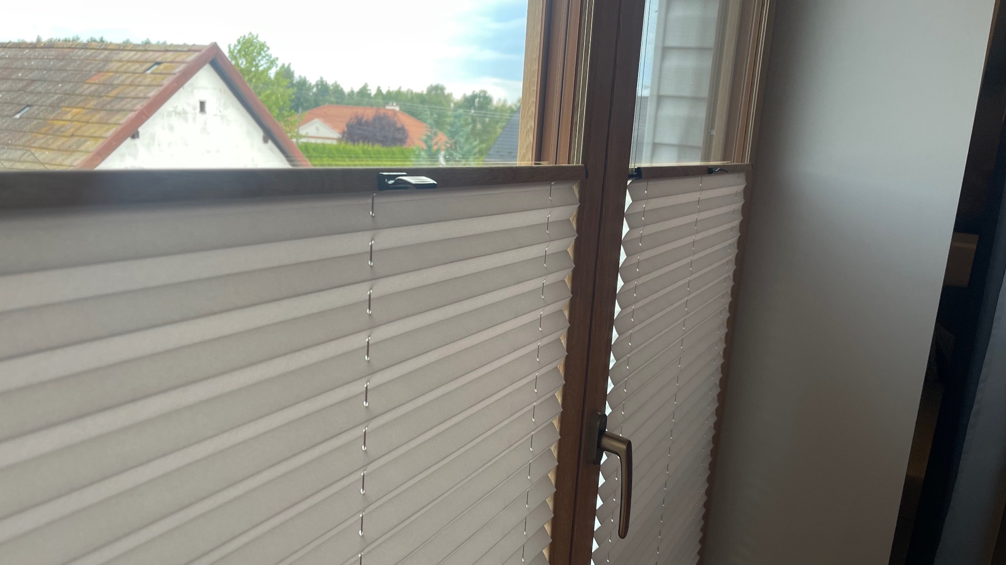 Zamknięte, białe, plisowane osłony okienne w drewnianej ramie, widok z wnętrza na domy w tle.