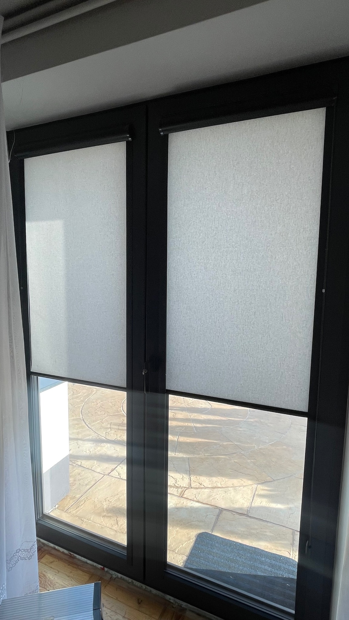 Dwa okna z czarnymi ramami wyposażone w jasne, teksturowane osłony przeciwsłoneczne, częściowo opuszczone, z widokiem na taras z kamienną nawierzchnią.