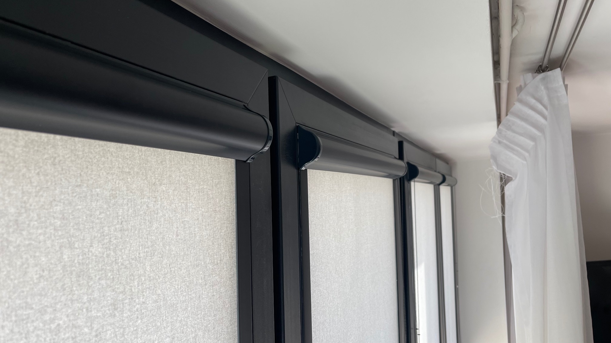 Fragment okna z zamontowanymi jasnymi roletami w czarnych kasetach, widok z dołu, z boku zwisająca biała firanka.
