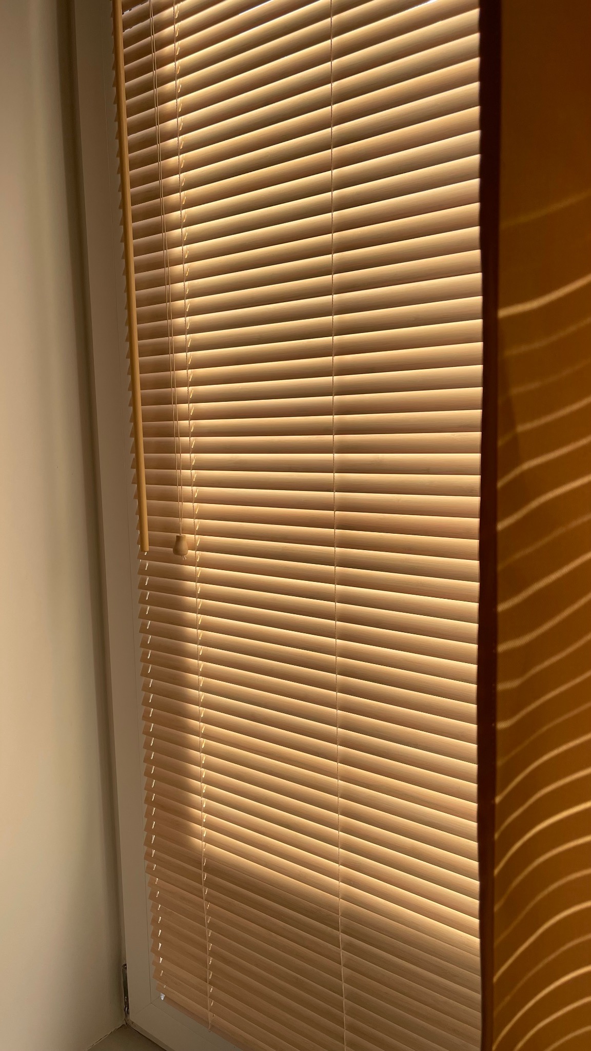 Zamknięte poziome żaluzje w odcieniach beżu, z widocznym sznurkiem sterującym po lewej stronie i fragmentem ściany w ciepłym świetle po prawej.