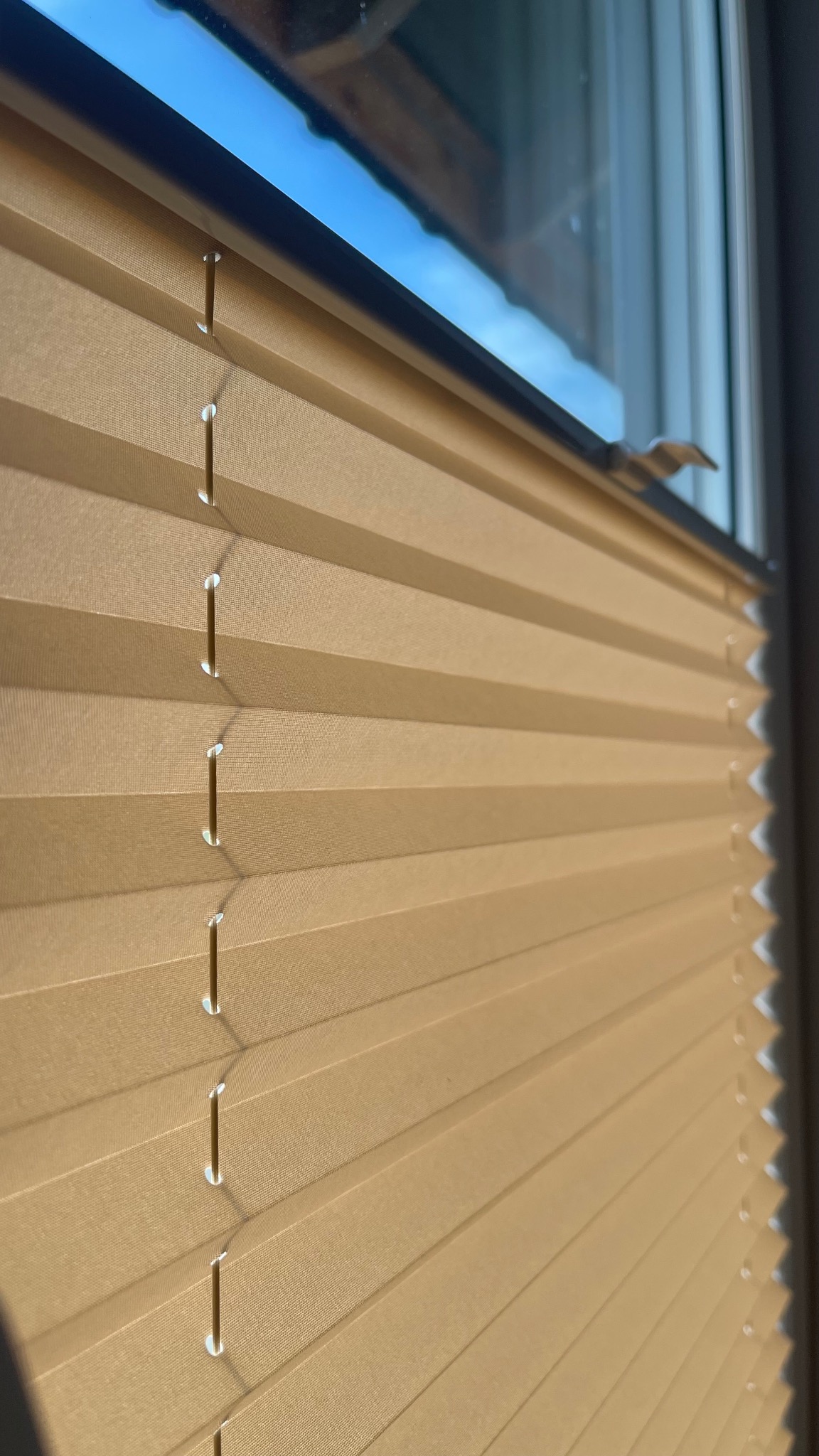 Beżowe, plisowane osłony okienne w pozycji zamkniętej, widoczne sznurki i fragment okna z odbijającym się niebem.