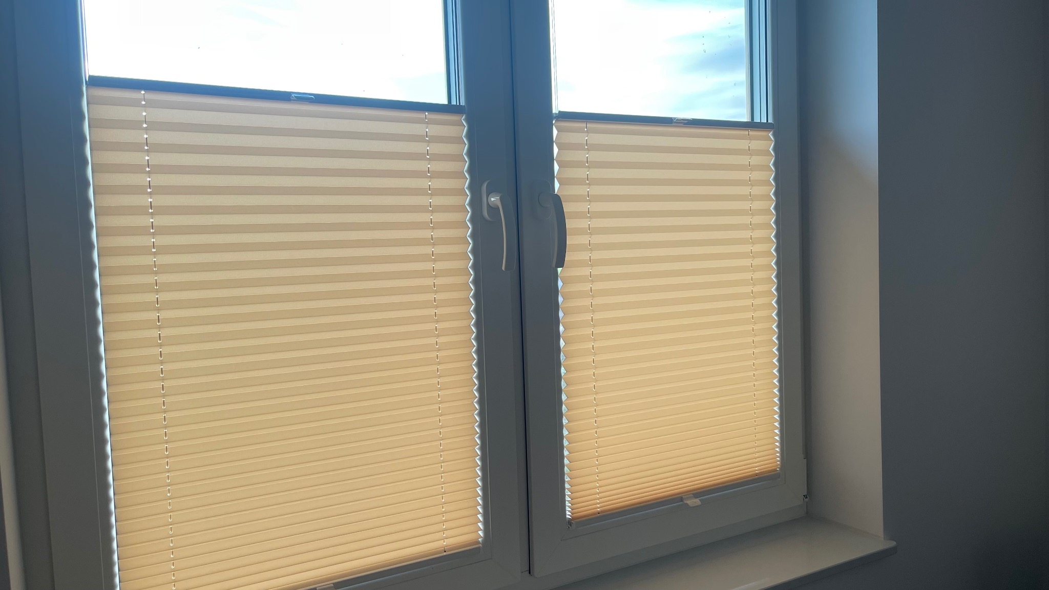 Beżowe, plisowane osłony okienne zamontowane w dwuskrzydłowym oknie PCV, widoczne z bliska.