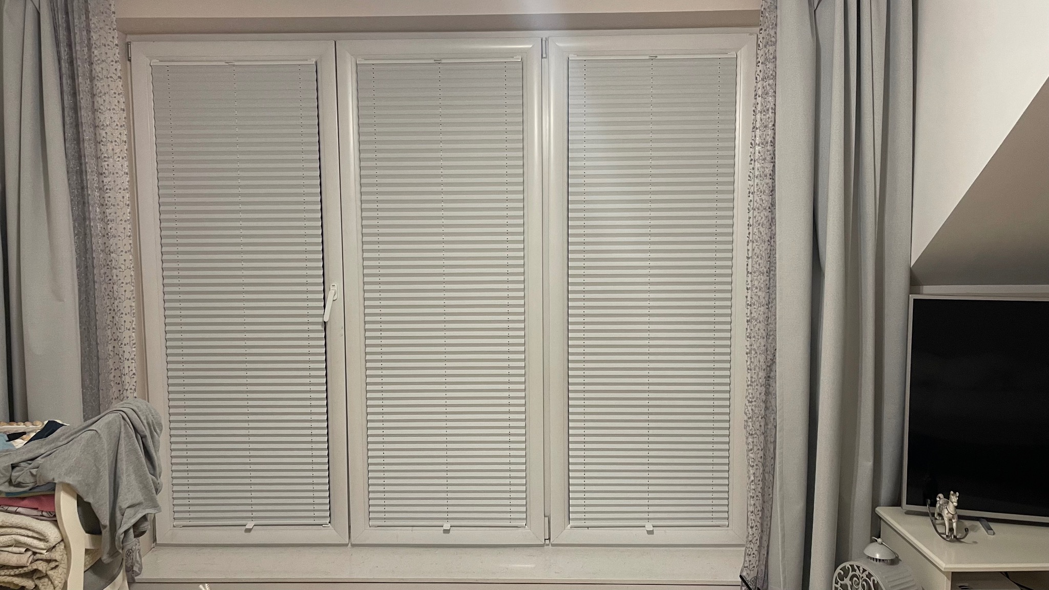 Trzy okna z zamontowanymi białymi, plisowanymi osłonami okiennymi, zasłonięte częściowo przez wzorzyste i gładkie zasłony. Widoczny fragment telewizora i dekoracje na komodzie.