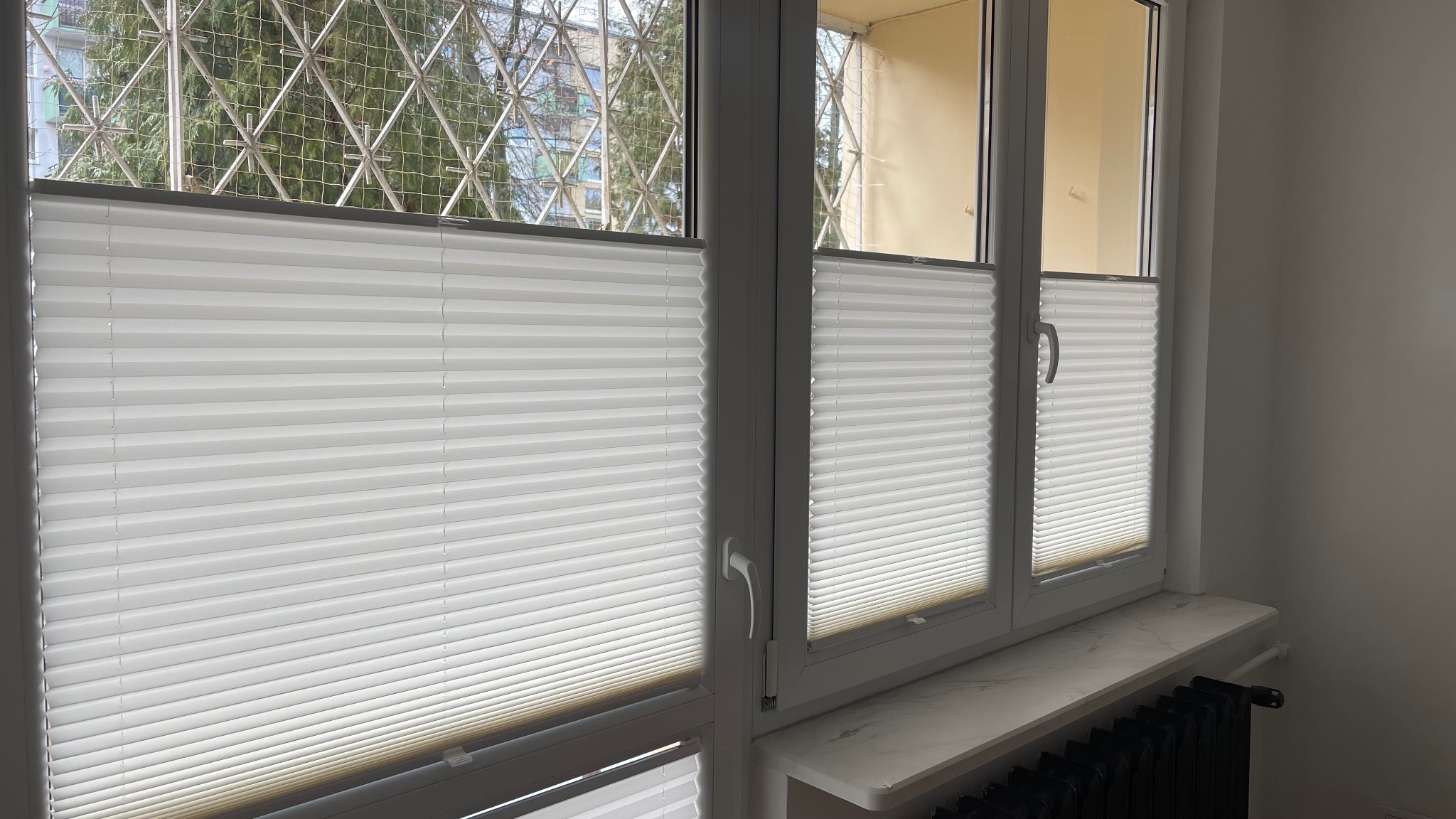 Białe, plisowane osłony okienne zamontowane w oknach z widokiem na drzewa za metalową kratą. Na parapecie imitacja marmuru, pod spodem czarny grzejnik.