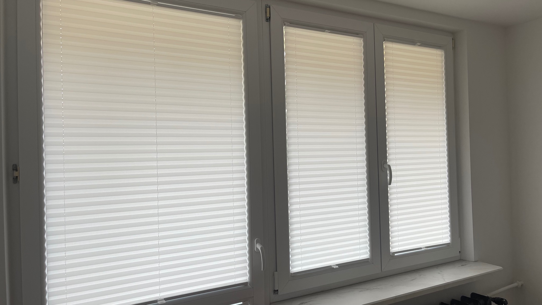 Trzy okna z zamontowanymi białymi plisami harmonijkowymi, widoczne detale mechanizmów sterujących.