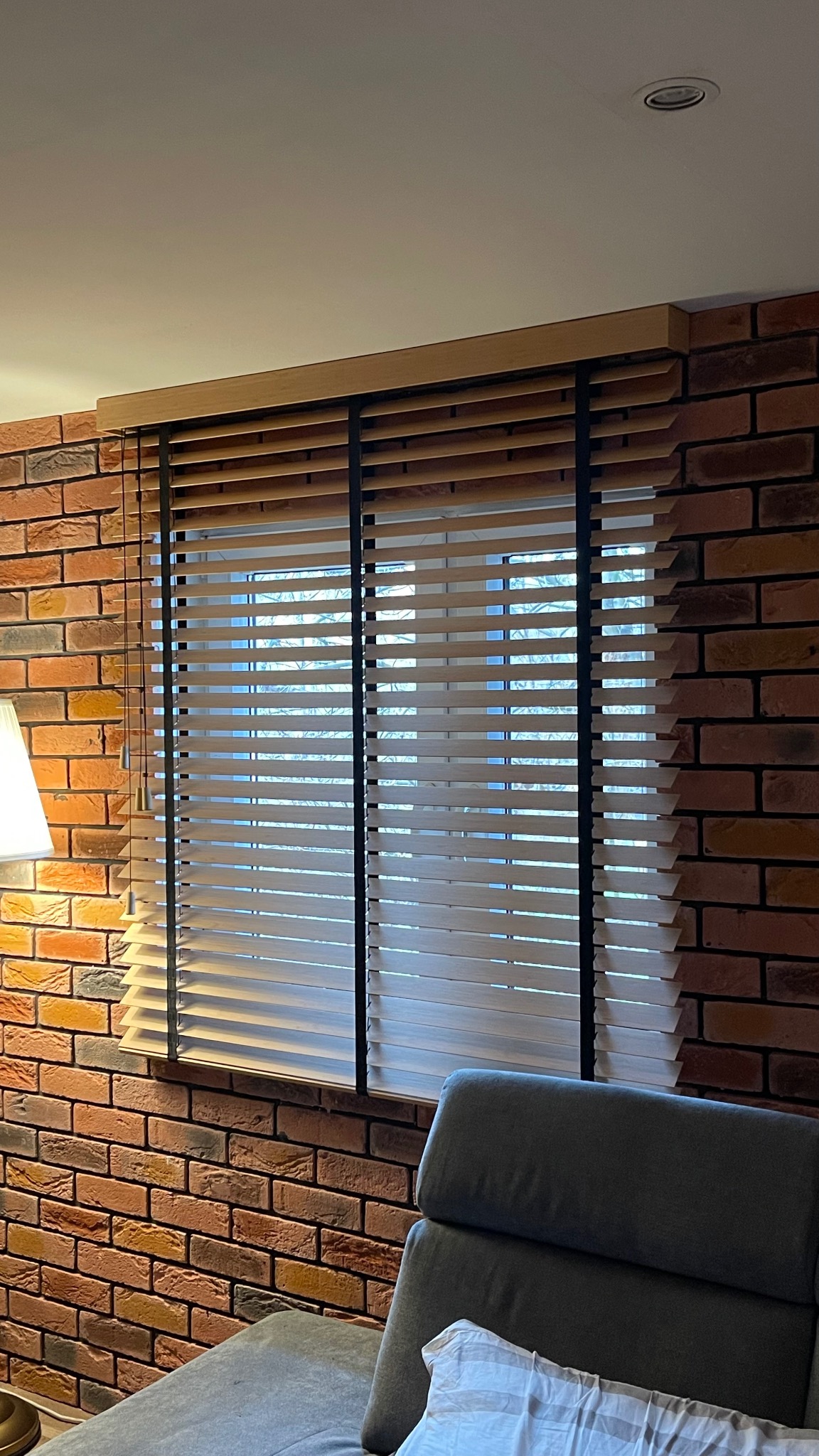 Drewniane żaluzje z czarnymi taśmami na tle ceglanej ściany, widoczne z poziomu szarej sofy.