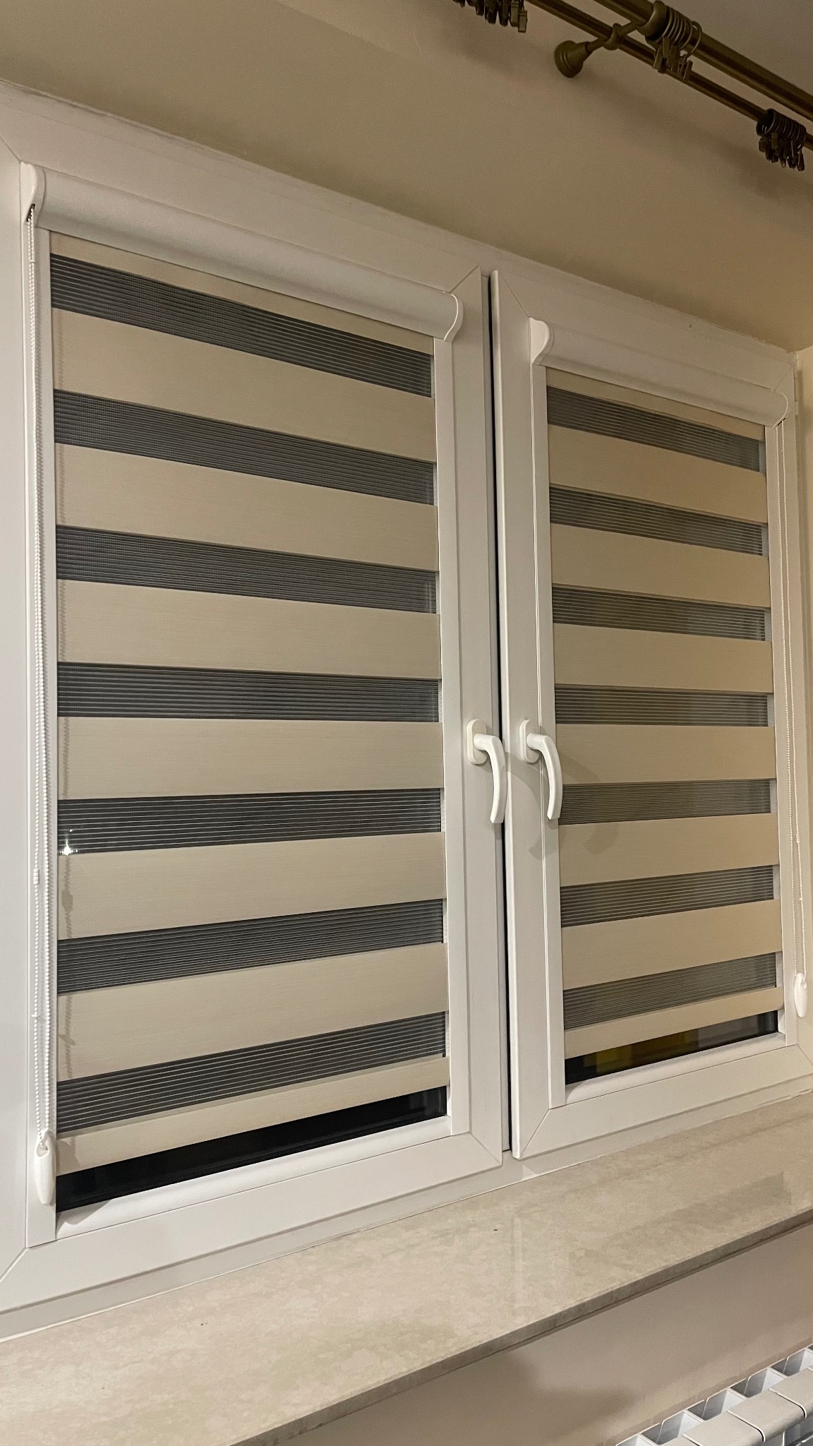 Dwuskrzydłowe okno z zamontowanymi roletami w biało-szare pasy, widoczne białe ramy okienne i parapet z jasnego kamienia.