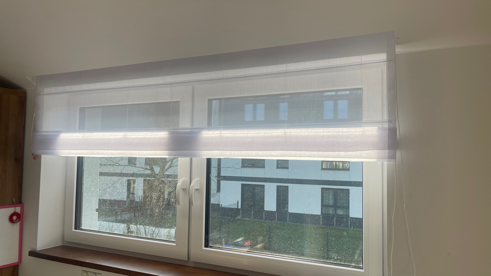 Delikatna, biała osłona okienna z materiału o subtelnej fakturze, częściowo podciągnięta, z widocznymi fałdami, na tle okna z widokiem na nowoczesny budynek.