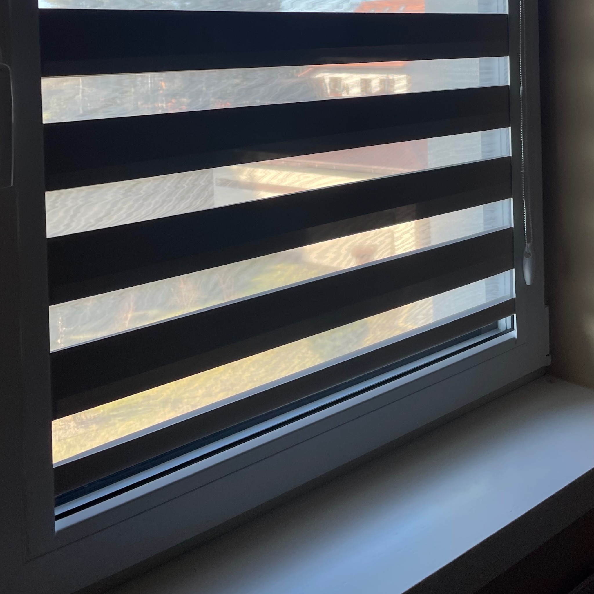 Okno z zamontowaną przesłoną okienną z poziomymi pasami zaciemniającymi i przepuszczającymi światło, widok na zewnątrz z delikatnym rozproszeniem światła.