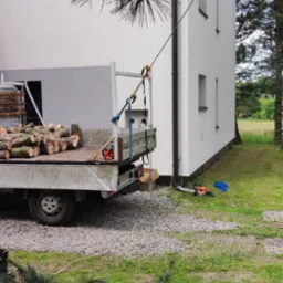 Przyczepa z pociętymi pniami drzewa na tle domu. Lina przymocowana do dachu. Obok leży piła motorowa. Widoczne gałęzie drzewa.