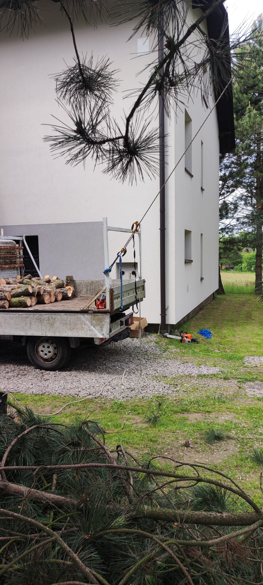 Przyczepa z pociętymi pniami drzewa na tle domu. Lina przymocowana do dachu. Obok leży piła motorowa. Widoczne gałęzie drzewa.