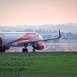 NotForSquares Arkadiusz Krzysztof - Samolot EasyJet startujący z lotniska, widok z tyłu, w tle zalesione wzgórza i delikatne różowe niebo o świcie.