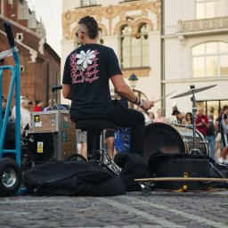 NotForSquares Arkadiusz Krzysztof - Uliczny perkusista gra na zestawie perkusyjnym przed zgromadzonym tłumem na tle architektury Krakowa, widoczny wózek transportowy i deskorolka.