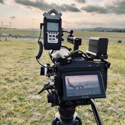 NotForSquares Arkadiusz Krzysztof - Profesjonalna kamera filmowa na statywie z podłączonym rejestratorem dźwięku Zoom H4n Pro, ustawiona na lotnisku podczas filmowania startującego samolotu, widocznego na ekranie kamery.