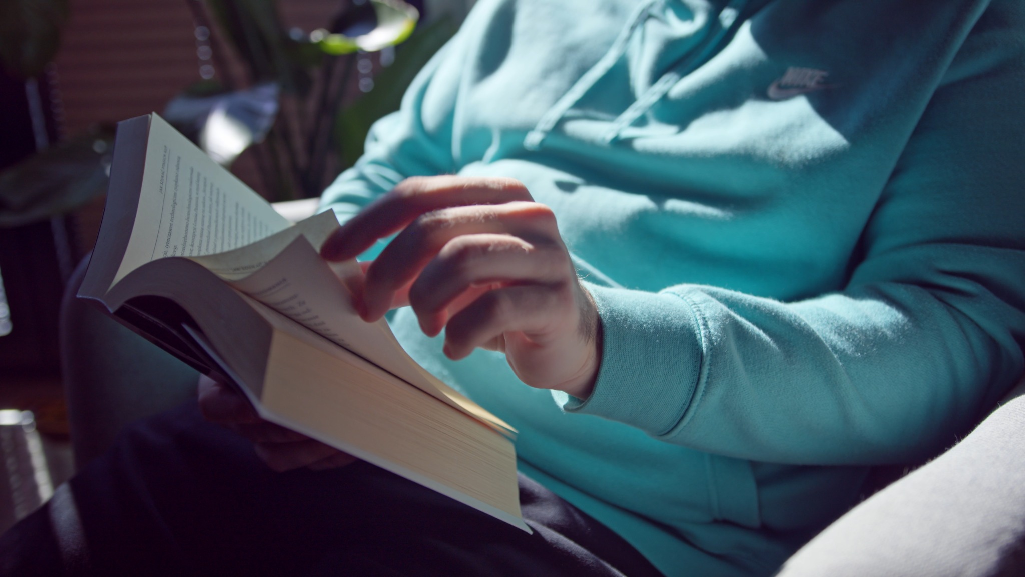Mężczyzna w błękitnej bluzie z kapturem czyta książkę w jasnym świetle słonecznym, widoczne dłonie przewracające strony.