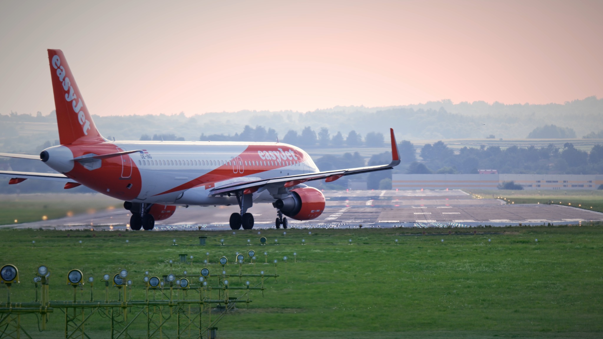 Samolot EasyJet startujący z lotniska, widok z tyłu, w tle zalesione wzgórza i delikatne różowe niebo o świcie.