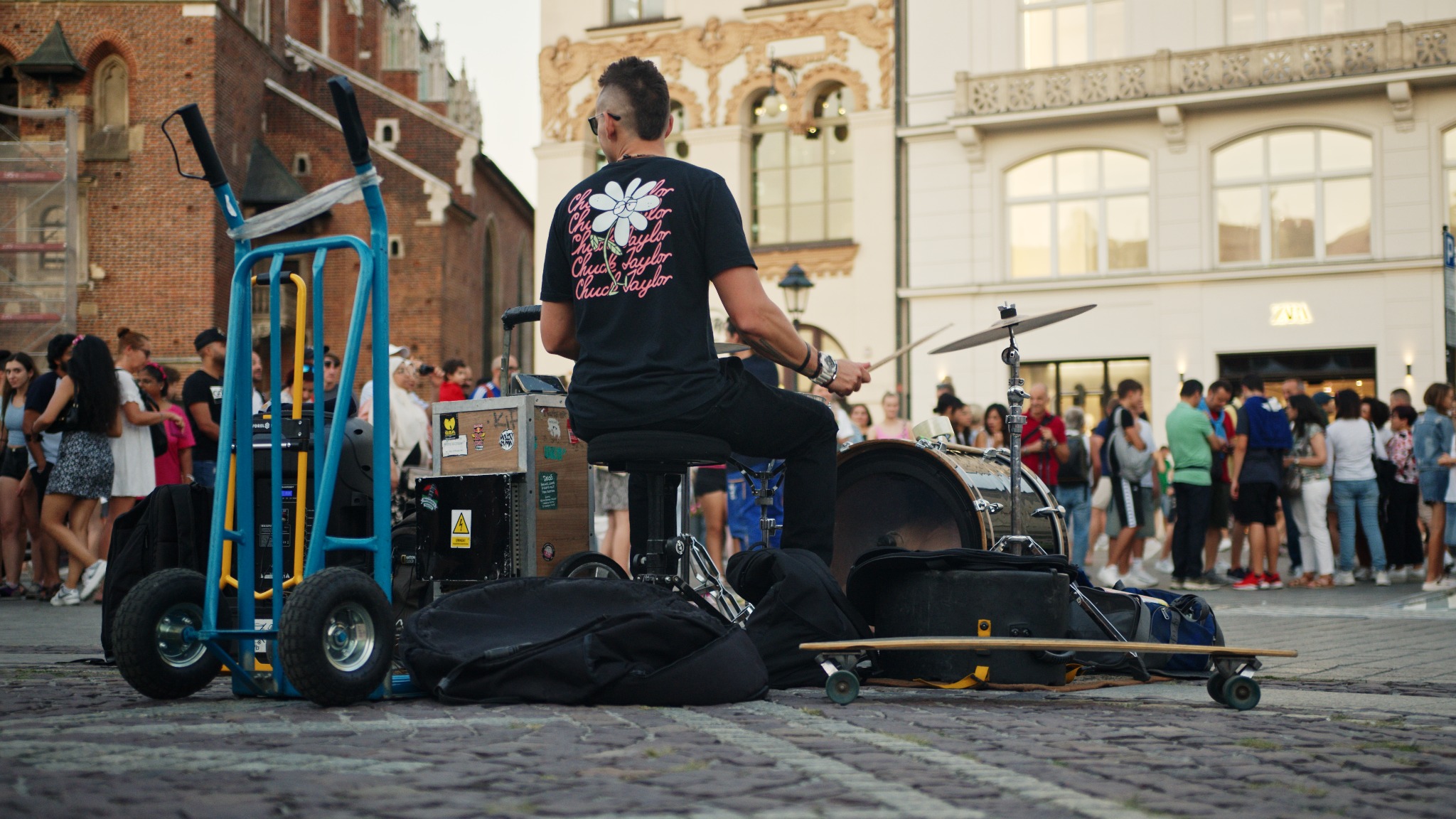 Uliczny perkusista gra na zestawie perkusyjnym przed zgromadzonym tłumem na tle architektury Krakowa, widoczny wózek transportowy i deskorolka.