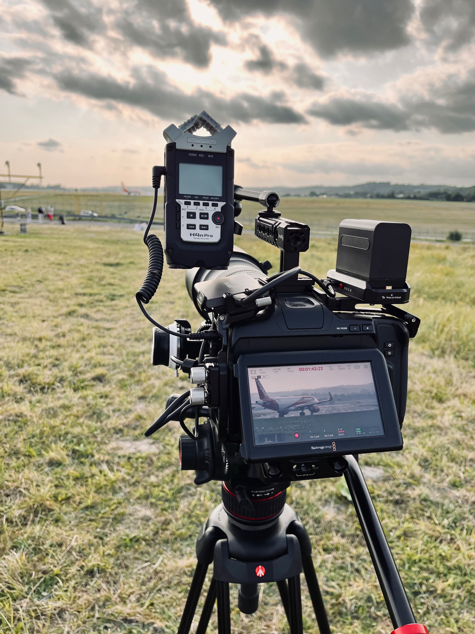 Profesjonalna kamera filmowa na statywie z podłączonym rejestratorem dźwięku Zoom H4n Pro, ustawiona na lotnisku podczas filmowania startującego samolotu, widocznego na ekranie kamery.