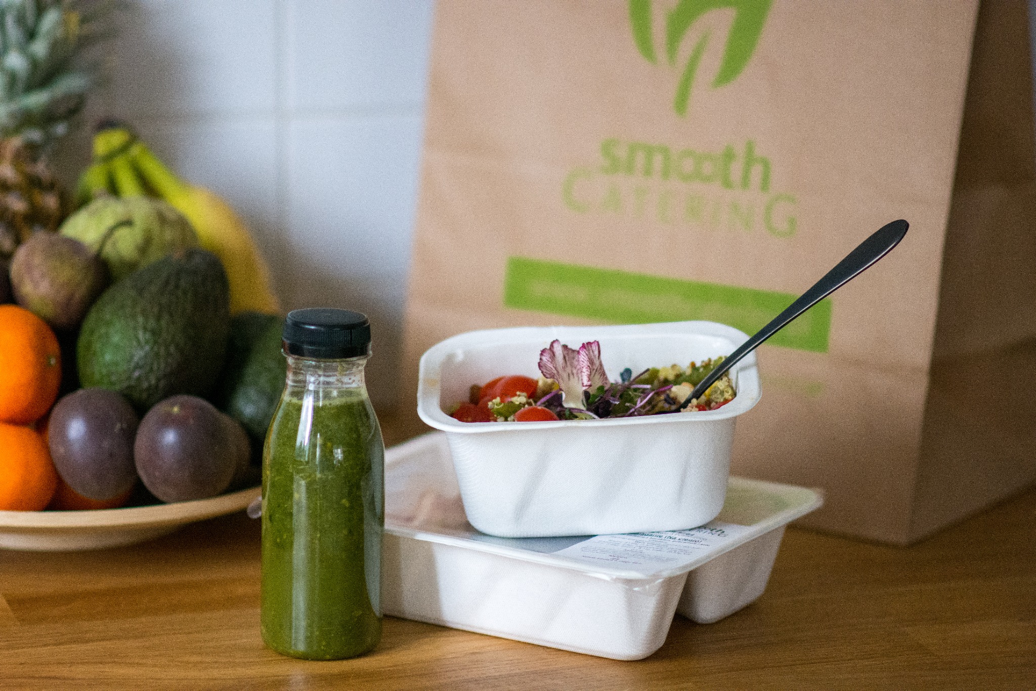 Zdjęcie prezentuje catering dietetyczny: butelka zielonego smoothie, sałatka z pomidorkami i kwiatem jadalnym w białym pojemniku, na tle papierowej torby z logo firmy cateringowej oraz talerza...