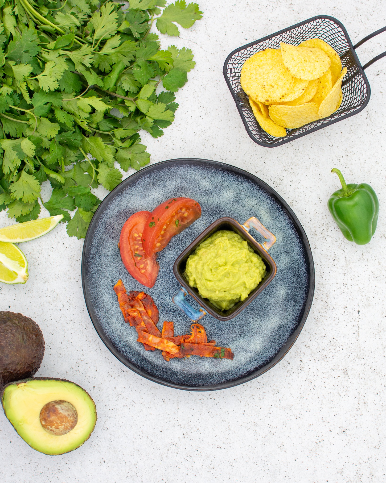 Kompozycja z guacamole w kwadratowej miseczce z uchwytami, chipsów w metalowym koszyku, plastrów pomidora, papryki i awokado na talerzu i obok niego, całość doświetlona z góry.