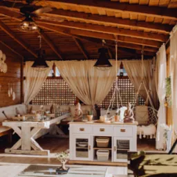 Drewniana altana z rustykalnym stołem, sofą z poduszkami, białymi zasłonami, dekoracjami makramowymi i figurkami Buddy, oświetlona ciepłym światłem.