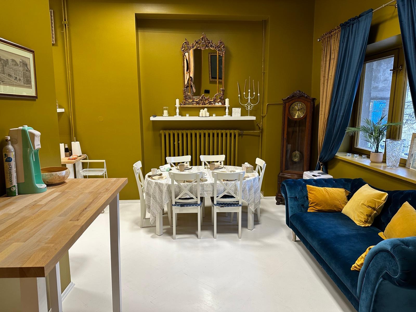 Ustawiony stół z białymi krzesłami w pokoju o oliwkowych ścianach, z dekoracyjnym lustrem, zegarem stojącym i niebieską sofą.