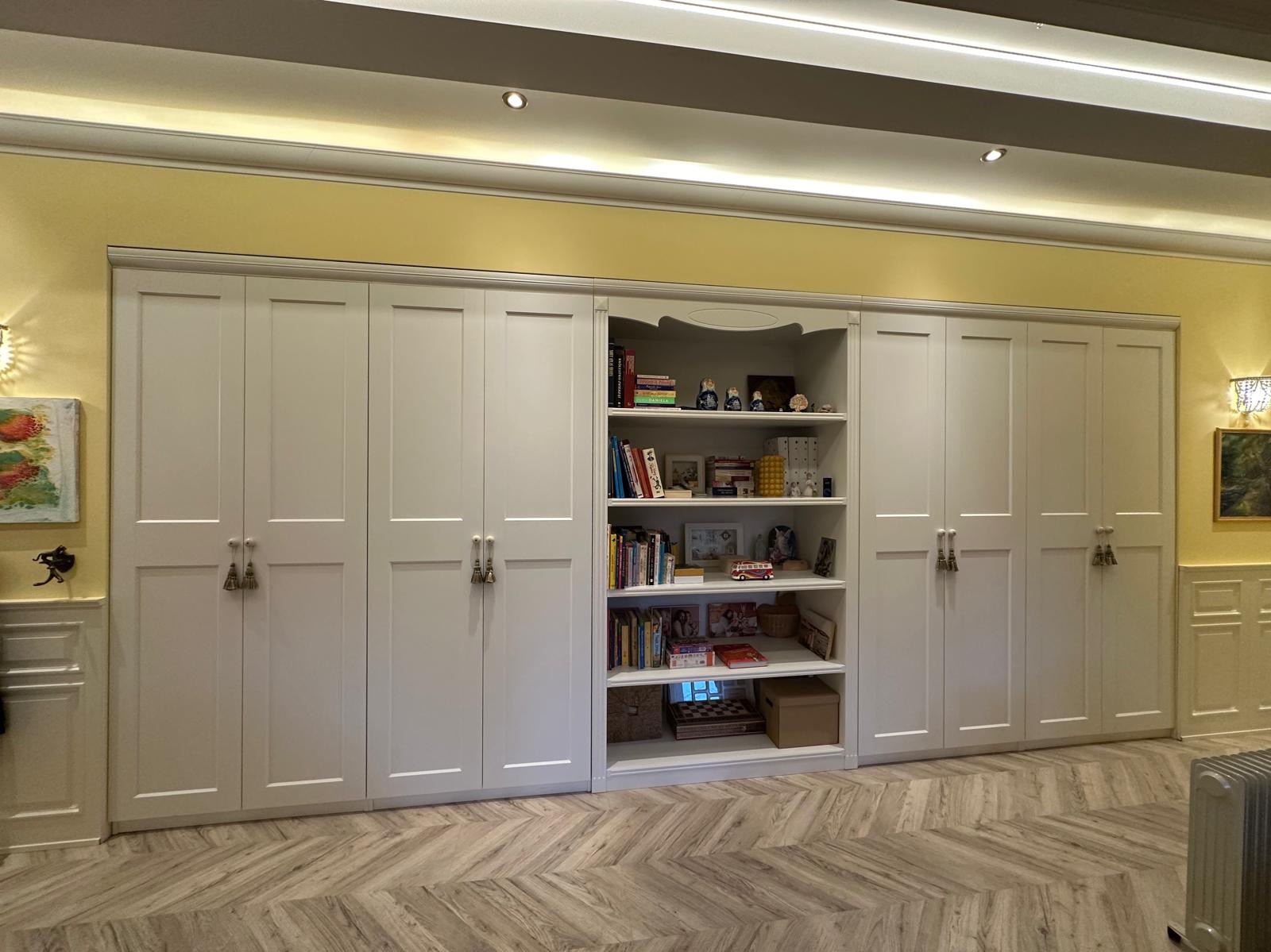 Biała zabudowa meblowa pod sufit z szafkami i regałem na książki, żółte ściany i parkiet w jodełkę.