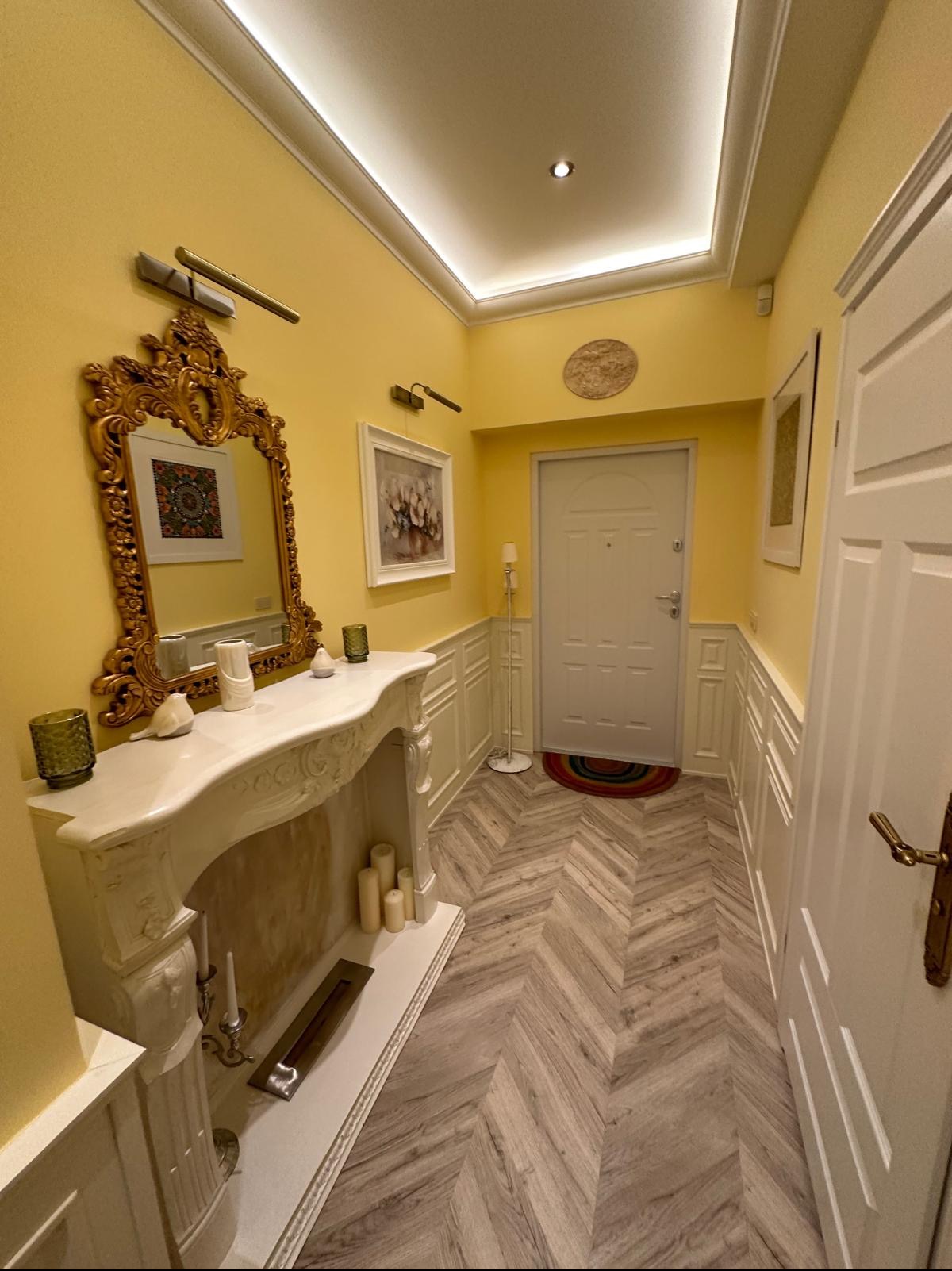 Jasny przedpokój z żółtymi ścianami, białymi panelami ściennymi, marmurowym kominkiem dekoracyjnym z lustrem w złotej ramie i podłogą ułożoną w jodełkę.