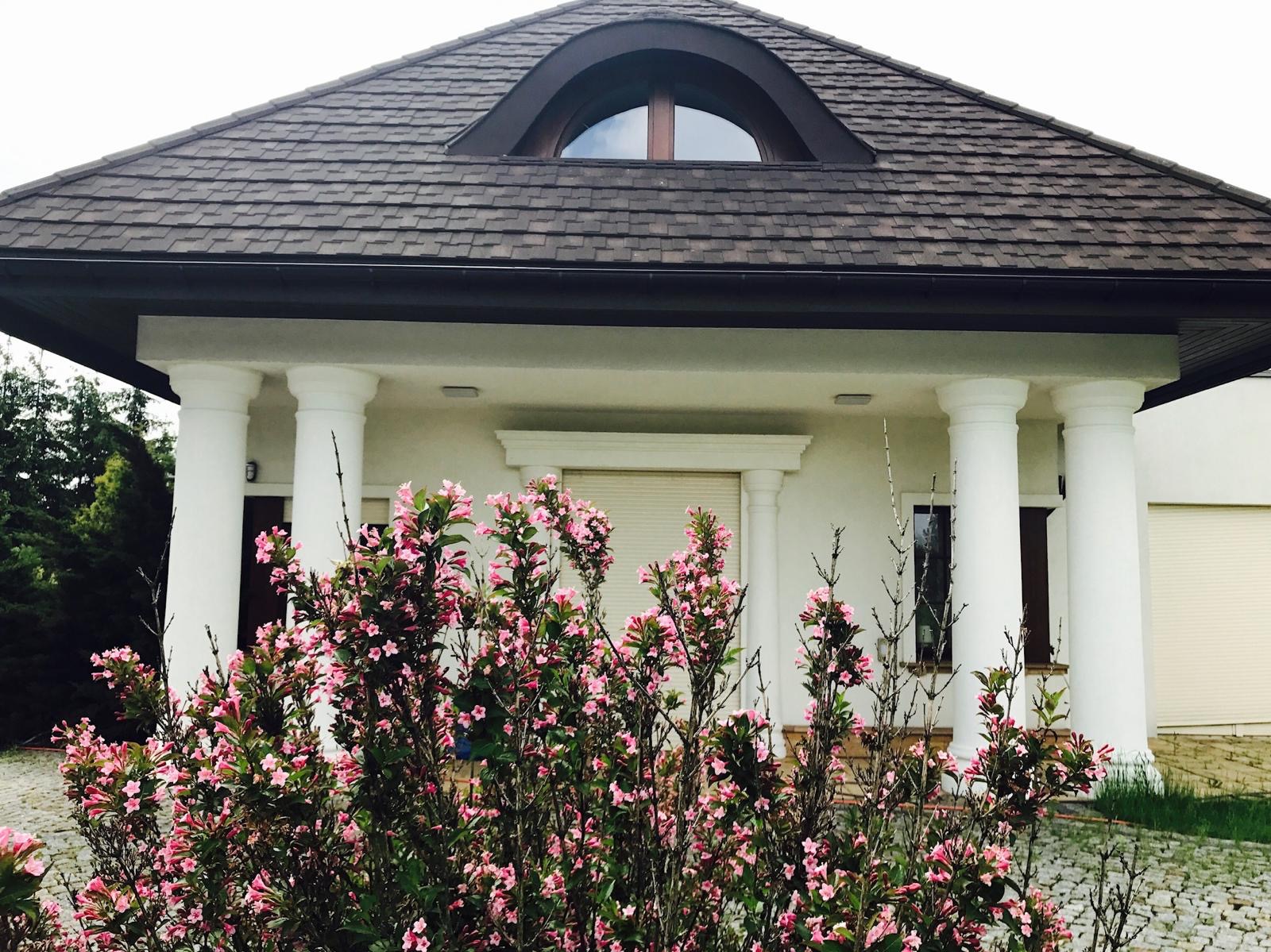 Elewacja domu jednorodzinnego z białymi kolumnami, brązowym dachem z gontów bitumicznych i oknem w kształcie półksiężyca, częściowo zasłonięta kwitnącym krzewem o różowych kwiatach.