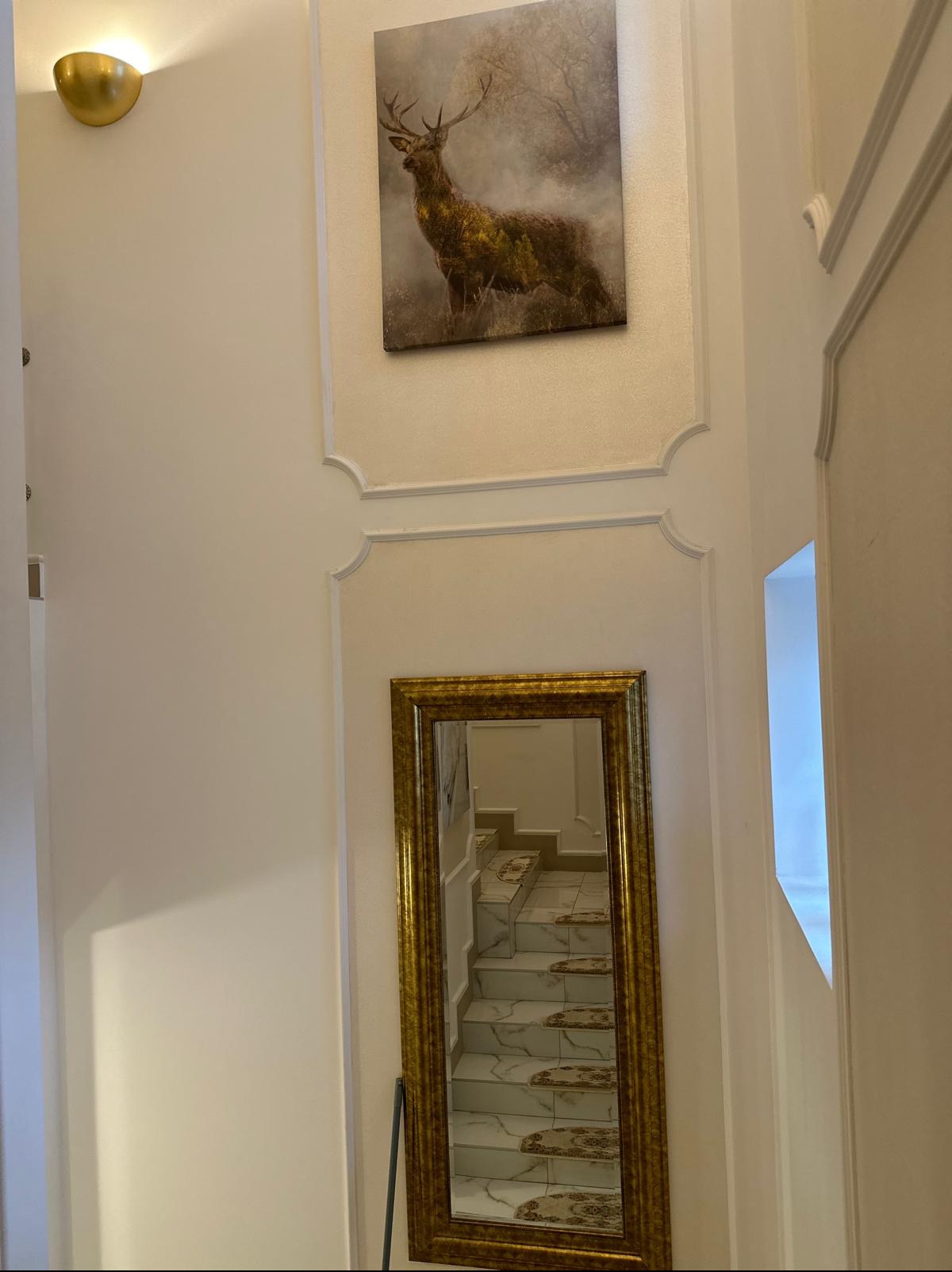 Klatka schodowa w jasnej tonacji z dekoracyjnymi listwami, lustrem w złotej ramie odbijającym schody i obrazem jelenia na ścianie.
