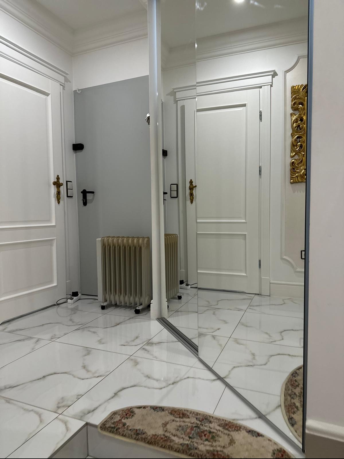 Wykończone wnętrze łazienki z białymi drzwiami z ozdobnymi klamkami, grzejnikiem żeberkowym i lustrem tworzącym iluzję przestrzeni, białe płytki podłogowe z szarymi żyłami.