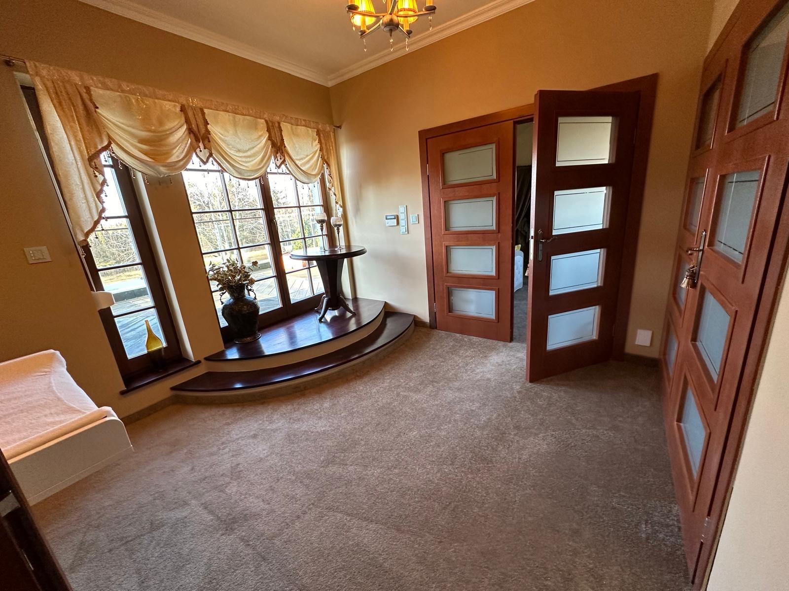 Przestronny przedpokój z dużym oknem, dekoracyjnymi zasłonami, ciemnym parkietem okalającym okno, okrągłym stolikiem, wazonem z kwiatami i drewnianymi drzwiami z przeszkleniami.