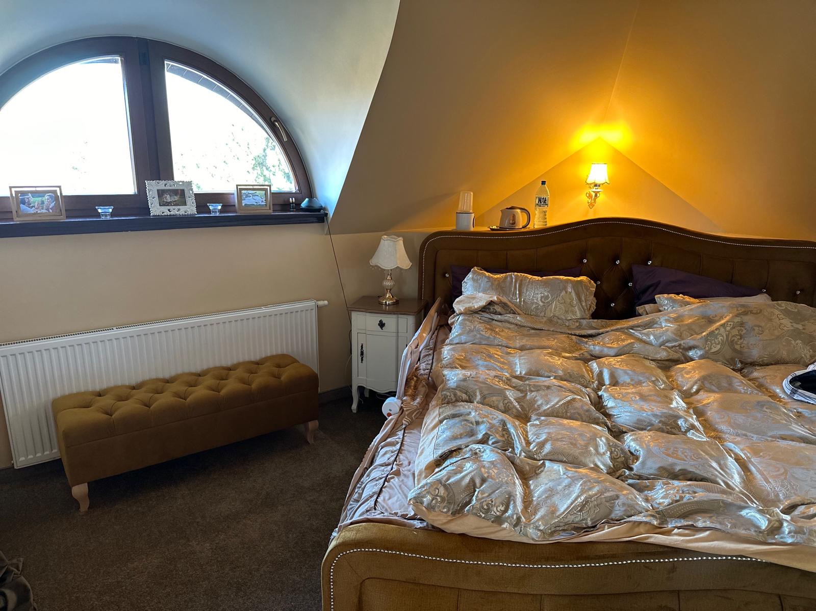 Sypialnia na poddaszu z oknem w kształcie łuku, brązowym łóżkiem z ozdobnym wezgłowiem, pikowaną pufą i białą szafką nocną.