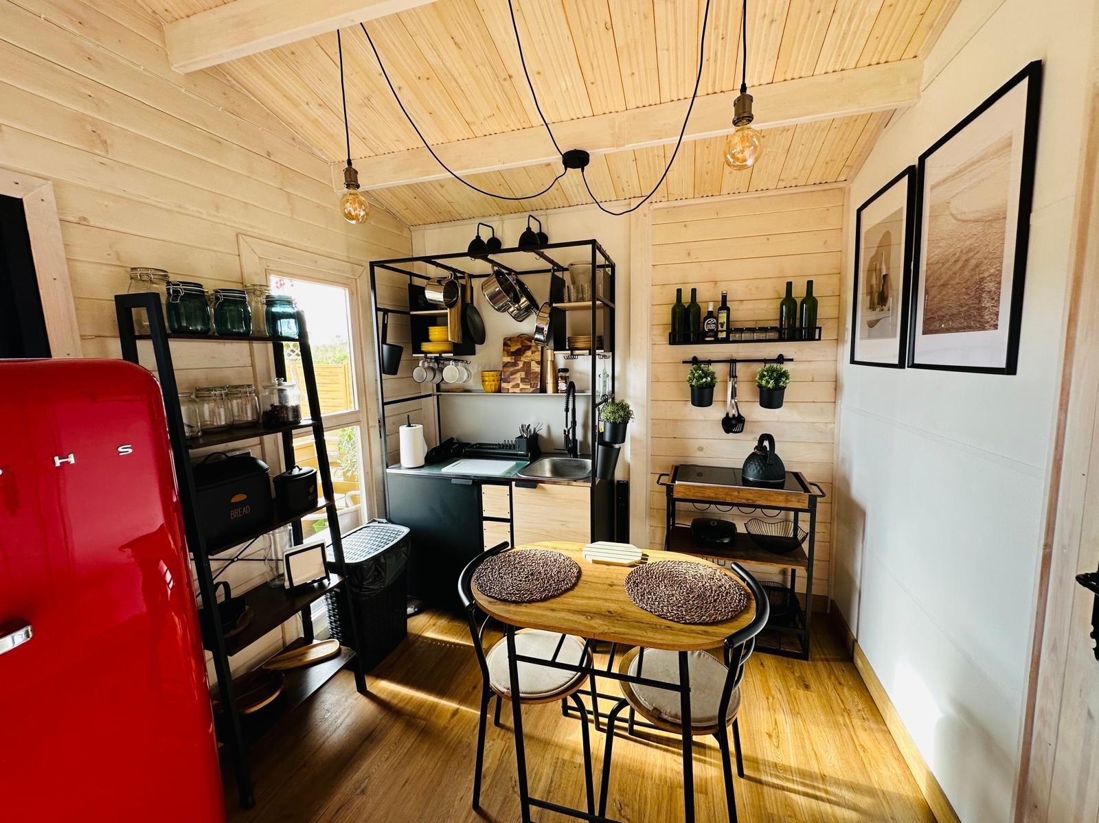 Jasne wnętrze domku z drewnianą boazerią, wyposażone w czerwoną lodówkę retro, czarne metalowe regały i stół z dwoma krzesłami, oświetlone żarówkami Edisona.