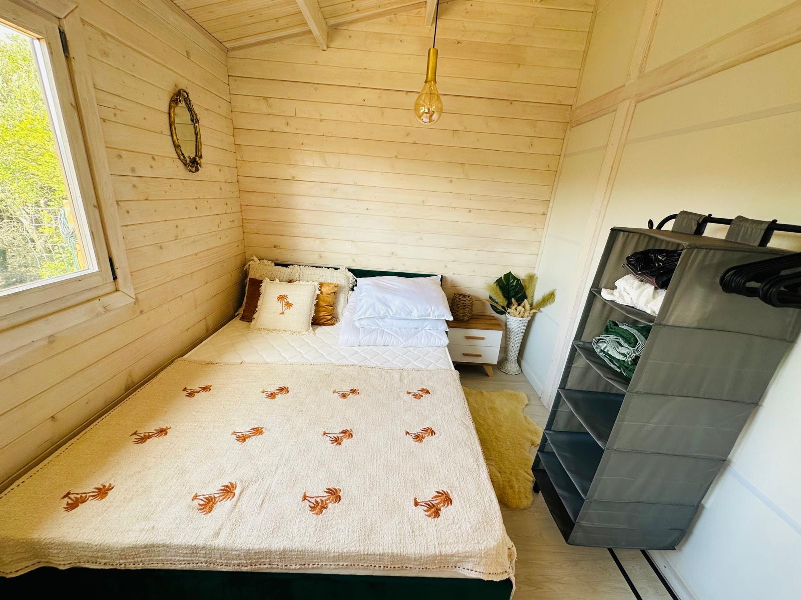 Wnętrze domku szkieletowego w Łodzi: sypialnia z drewnianymi ścianami, łóżkiem z narzutą w palmy, szafką nocną i wiszącą lampą, ukazująca przytulne wykończenie.