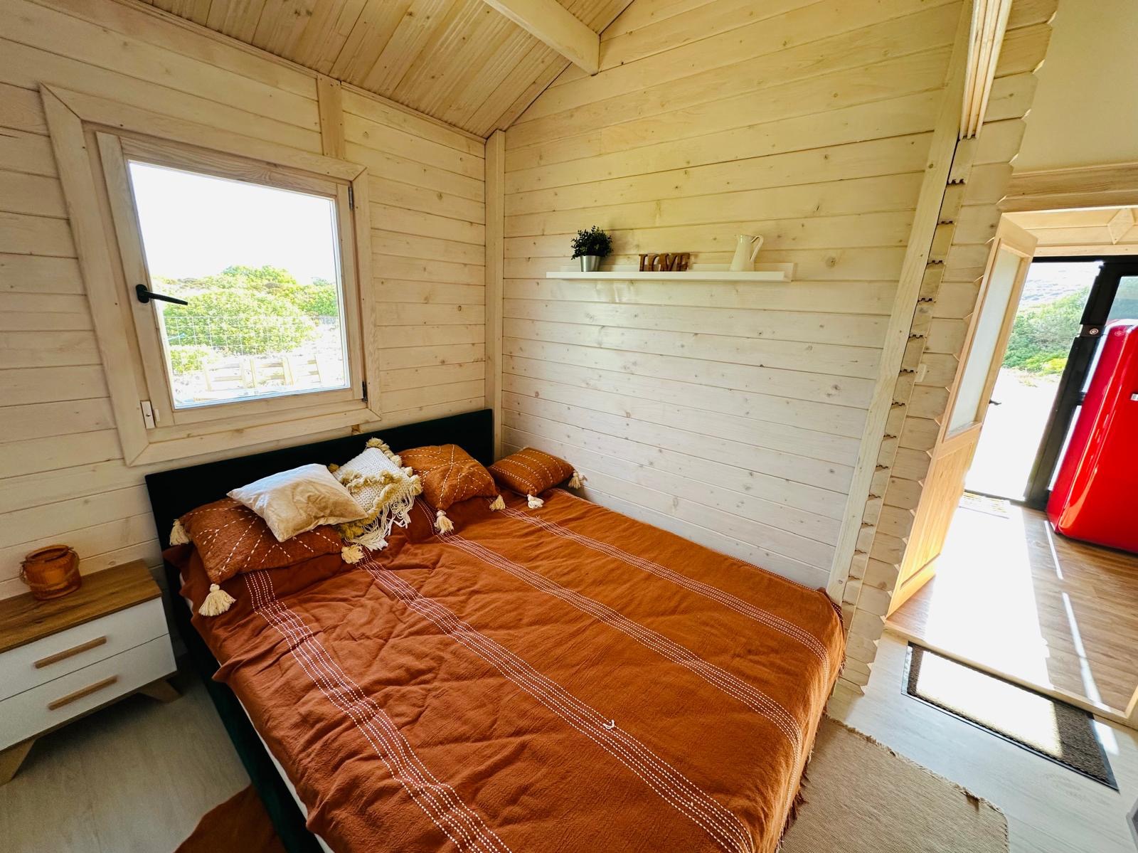 Wnętrze domku z drewnianymi ścianami, łóżkiem z pomarańczową narzutą i poduszkami w stylu boho, okno z widokiem na zewnątrz, półka z dekoracjami i czerwona lodówka w tle.