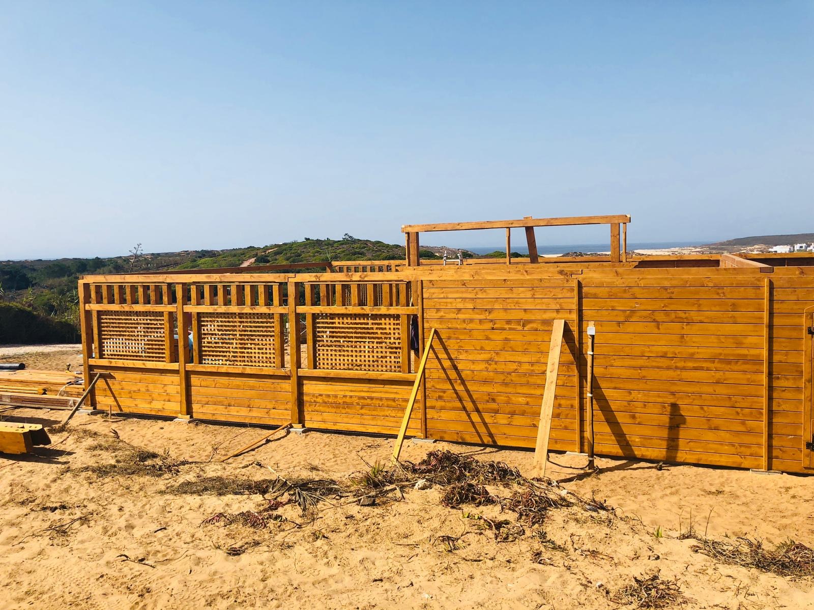 Konstrukcja z jasnego drewna, prawdopodobnie taras lub element domu szkieletowego, w trakcie budowy na piaszczystym terenie z widokiem na morze w oddali.