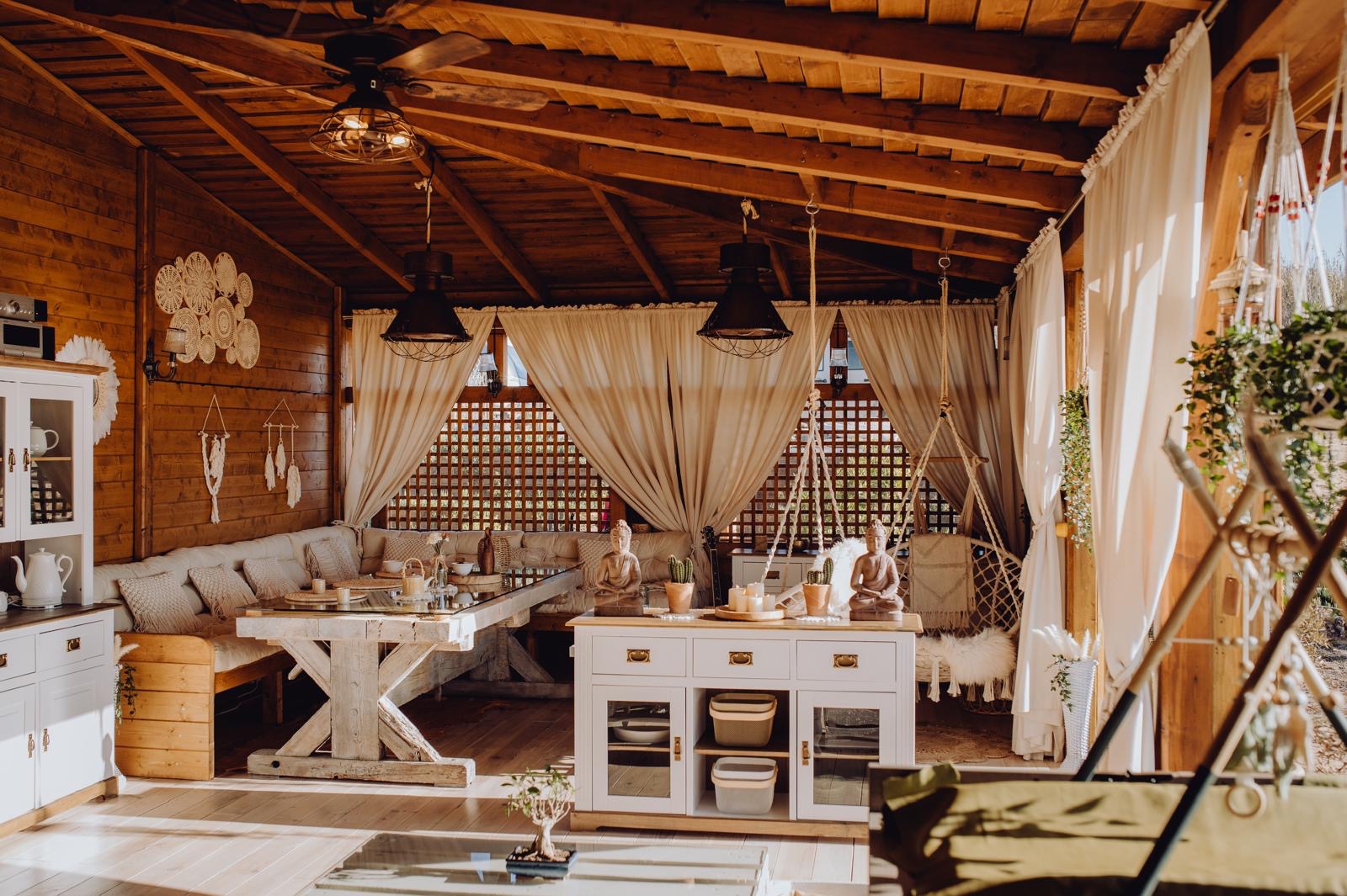 Drewniana altana z rustykalnym stołem, sofą z poduszkami, białymi zasłonami, dekoracjami makramowymi i figurkami Buddy, oświetlona ciepłym światłem.