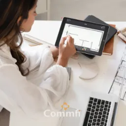 Kobieta w białej koszuli rysuje plan mieszkania na tablecie za pomocą stylusa, widoczne biurko z laptopem, linijką, próbkami materiałów i planami architektonicznymi.