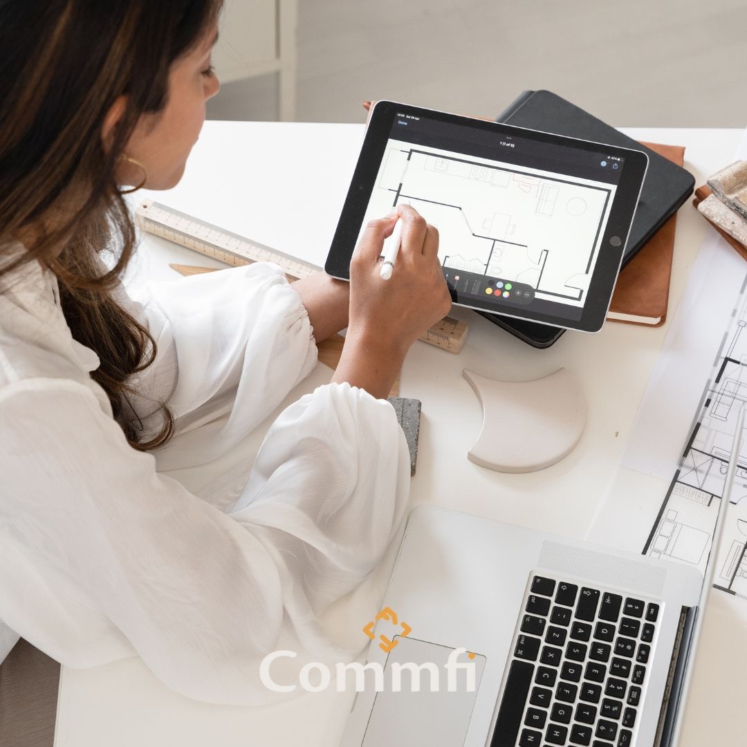 Kobieta w białej koszuli rysuje plan mieszkania na tablecie za pomocą stylusa, widoczne biurko z laptopem, linijką, próbkami materiałów i planami architektonicznymi.