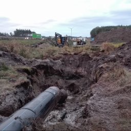 COBER s.c. - Rury ułożone w wykopie ziemnym, w tle maszyny budowlane i krajobraz z autostradą oraz turbiną wiatrową