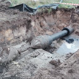 COBER s.c. - Wykop z widocznymi dwoma wiertłami do robót podziemnych, zanurzonymi w błocie i wodzie, z fragmentem rury w tle i taśmą ostrzegawczą u góry.