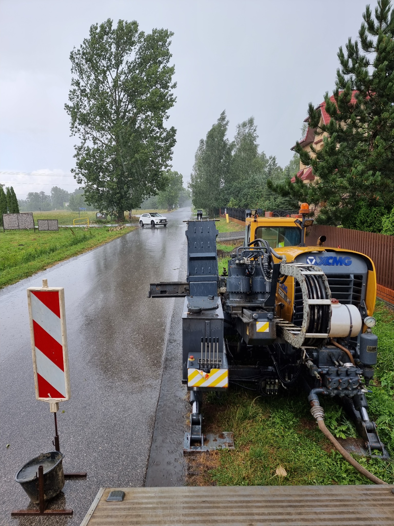 Żółto-szara maszyna do przewiertów sterowanych XCMG FZ200 w trakcie pracy na mokrej drodze, z widocznym znakiem ostrzegawczym i wiadrem obok, w tle biały samochód i drzewa w deszczowy dzień.