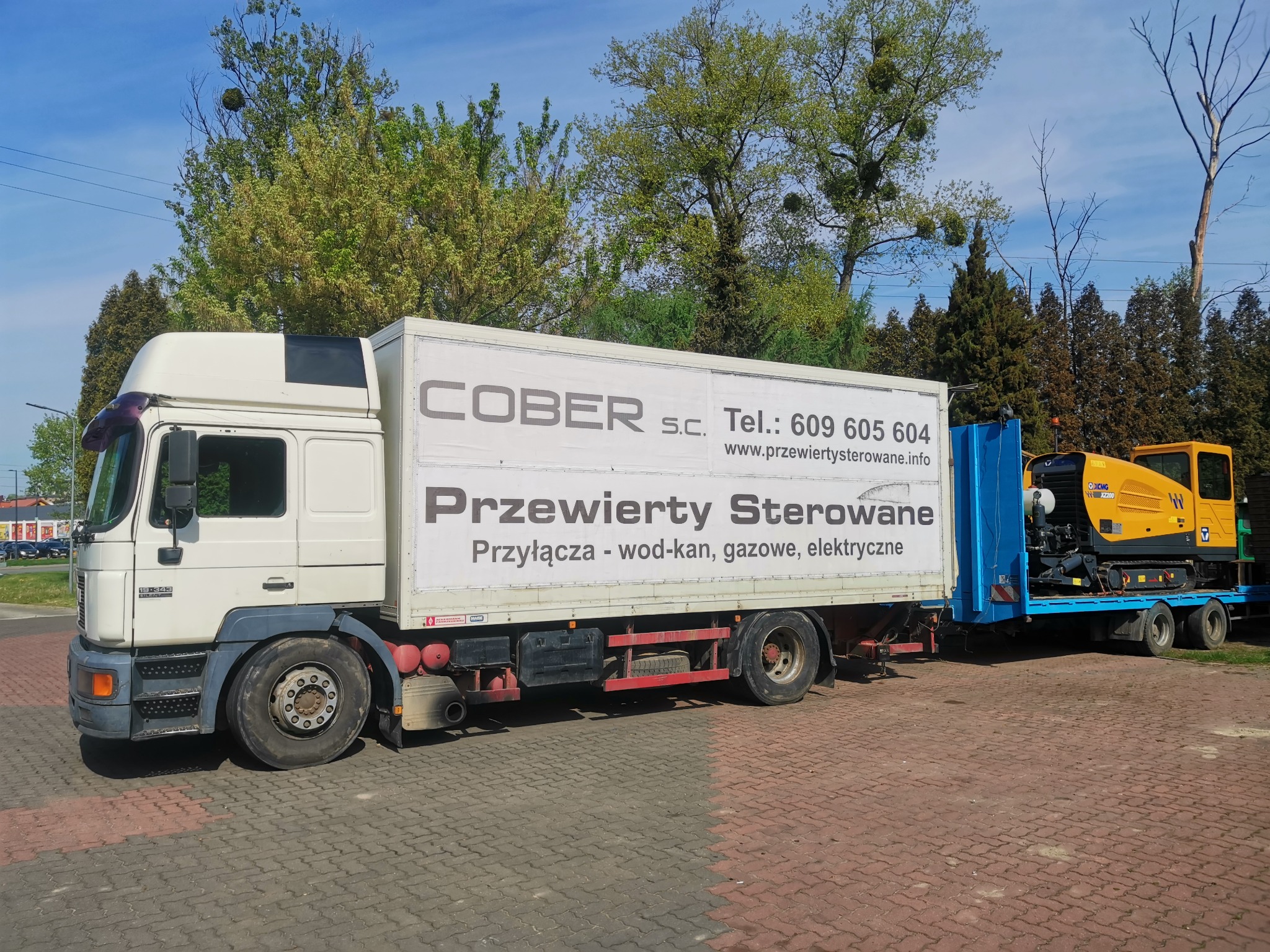 Ciężarówka z reklamą firmy COBER S.C. specjalizującej się w przyłączach wod-kan, gazowych i elektrycznych, z żółtą maszyną wiertniczą XCMG WX2200 na platformie z tyłu, zaparkowana na brukowanym placu.