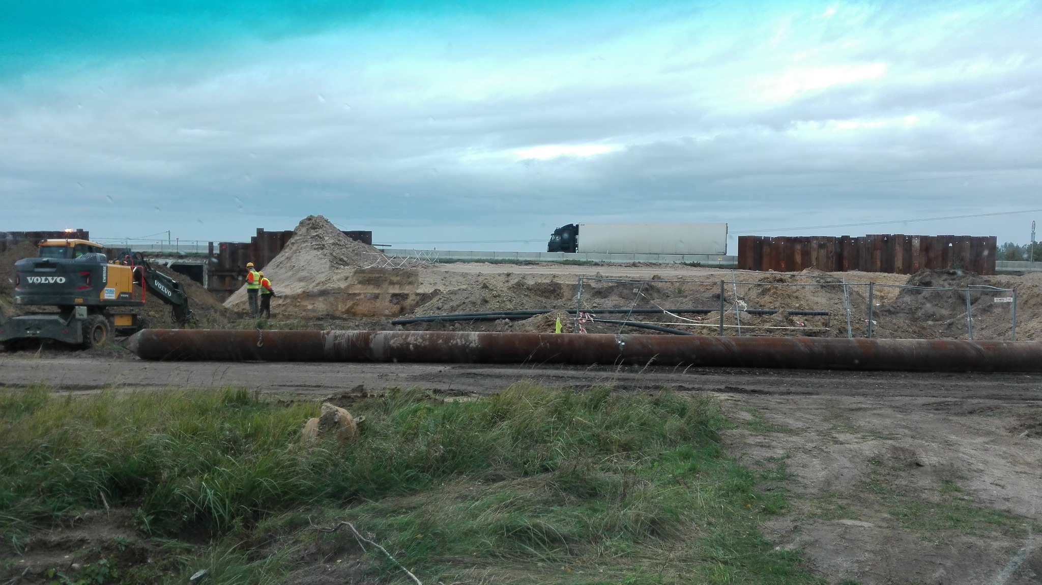 Budowa infrastruktury: Dwóch pracowników w odblaskowych kamizelkach konsultuje się obok koparki Volvo, z widocznymi rurami na pierwszym planie i ciężarówką w tle.