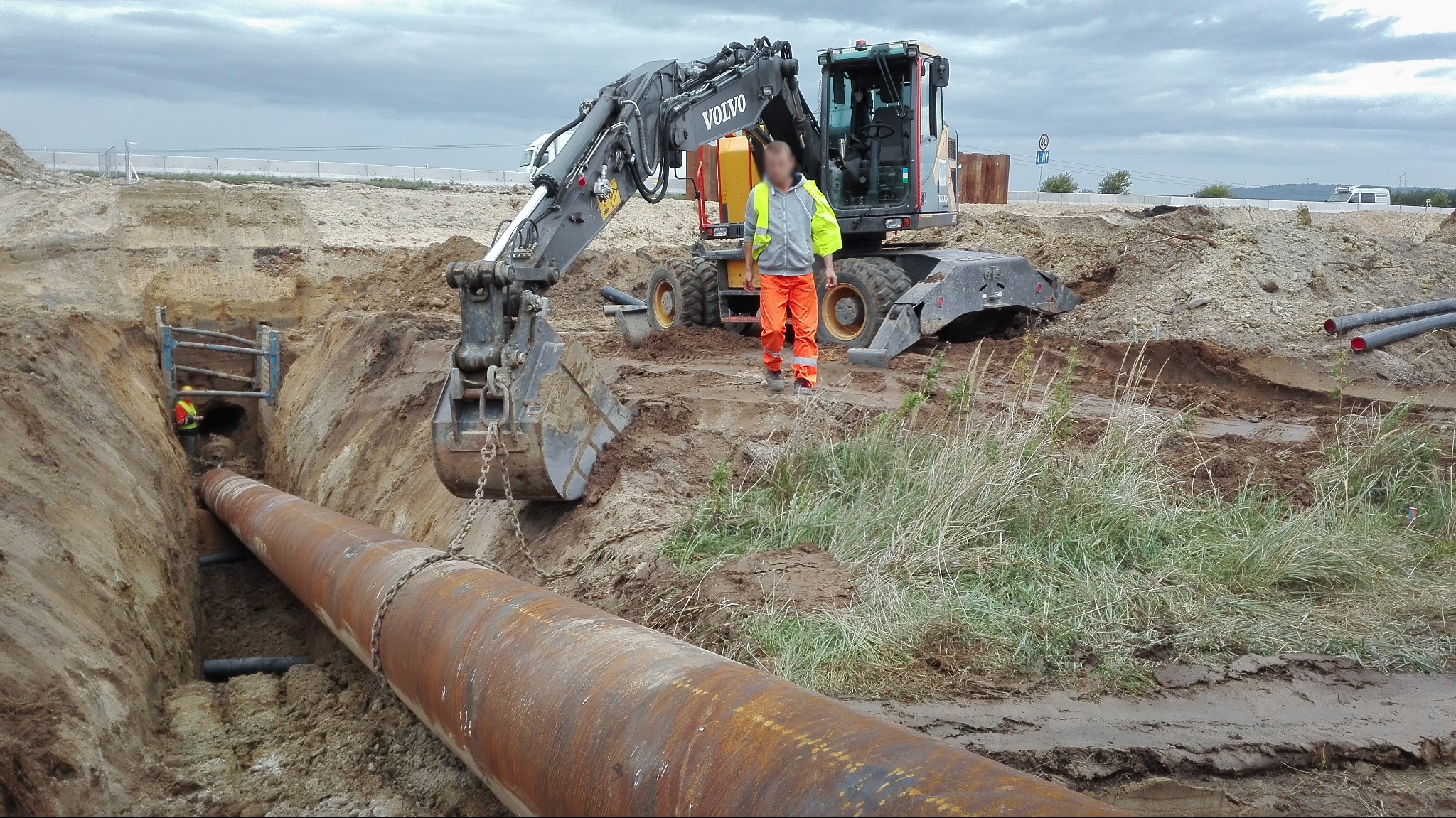 Układanie rury w wykopie z użyciem koparki Volvo; pracownik w pomarańczowych spodniach i żółtej kamizelce nadzoruje proces.