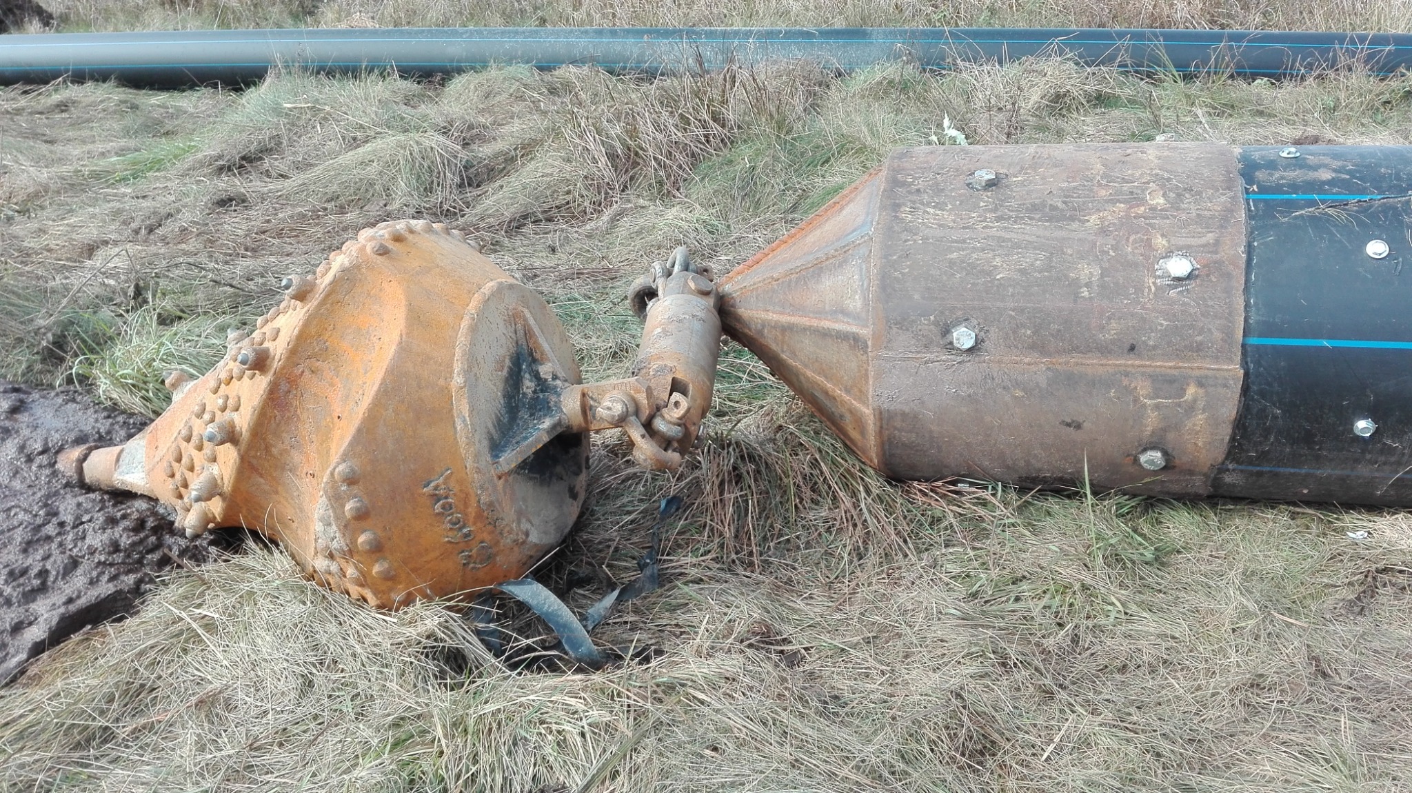 Zardzewiała głowica wiertnicza z licznymi wypustkami i fragment rury z niebieskim paskiem, leżące na wyschniętej trawie.