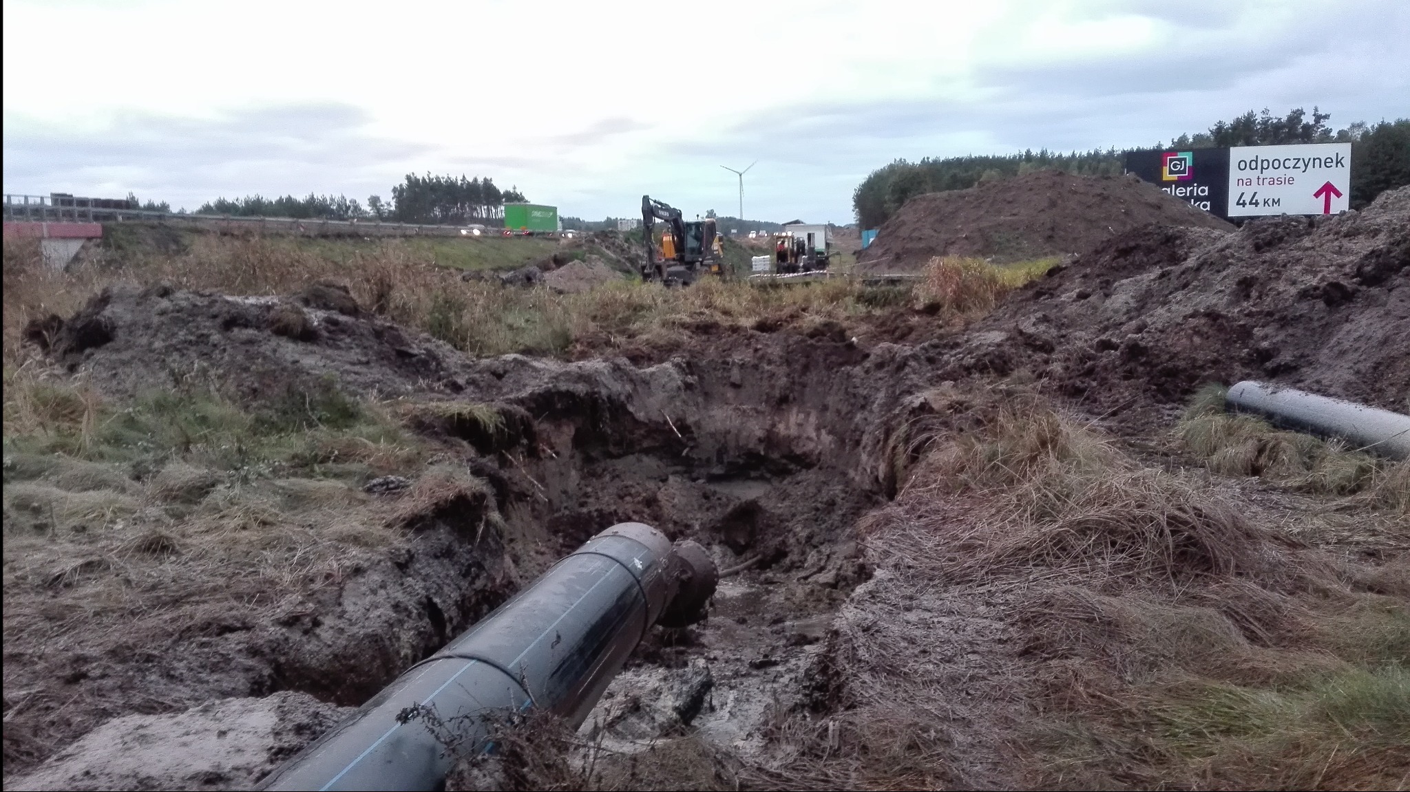 Rury ułożone w wykopie ziemnym, w tle maszyny budowlane i krajobraz z autostradą oraz turbiną wiatrową