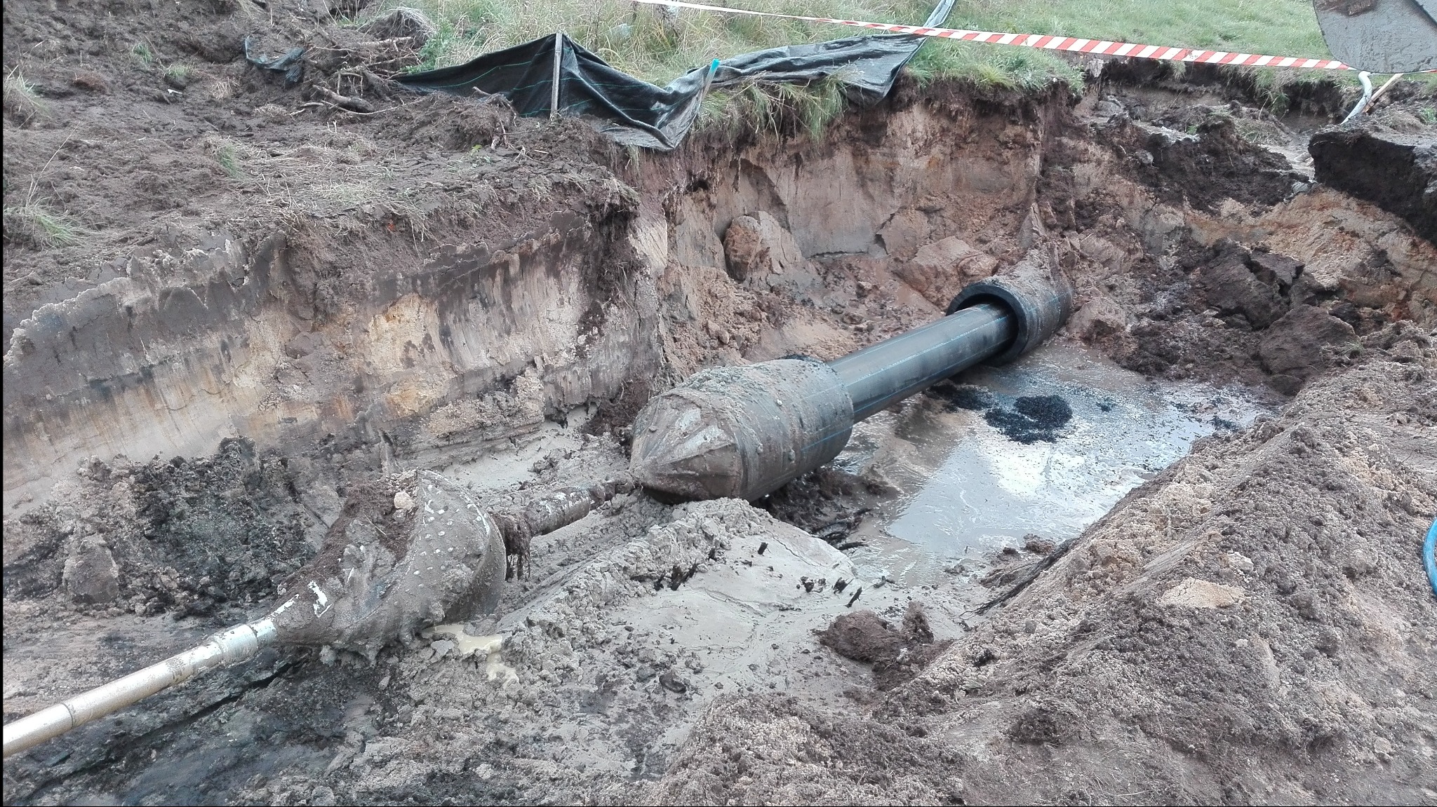 Wykop z widocznymi dwoma wiertłami do robót podziemnych, zanurzonymi w błocie i wodzie, z fragmentem rury w tle i taśmą ostrzegawczą u góry.
