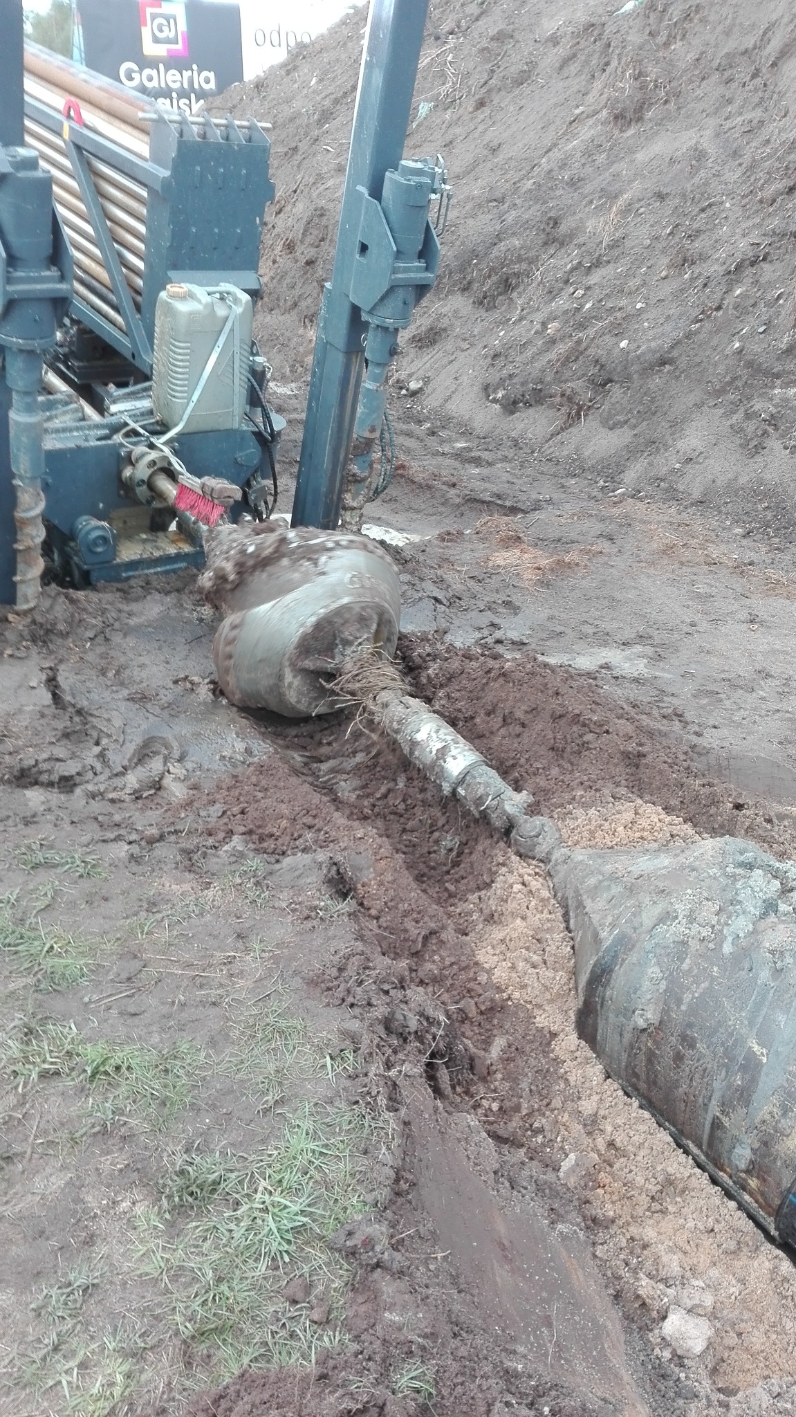 Przewiert sterowany w trakcie pracy: głowica wiertnicza zagłębiona w ziemi, widoczne błoto i wykop, maszyna wiertnicza w tle.